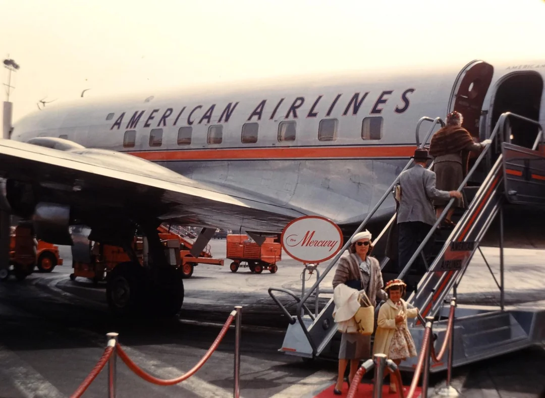 Passengers board an American Airlines plane using outdoor stairs. A woman and child in vintage clothing stand at the bottom, while others ascend. The scene appears to be from the mid-20th century.