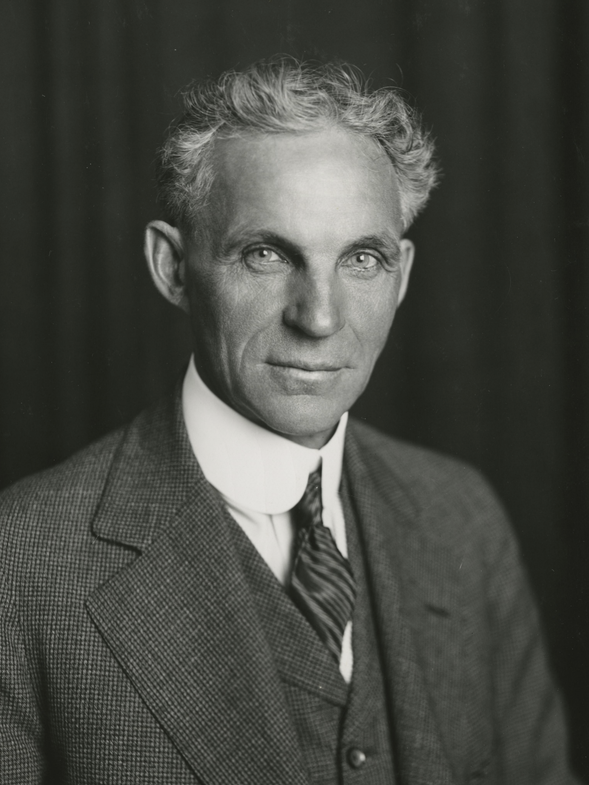 A black and white portrait of an older man with wavy gray hair, wearing a suit, vest, white shirt, and tie, posing in front of a dark backdrop.