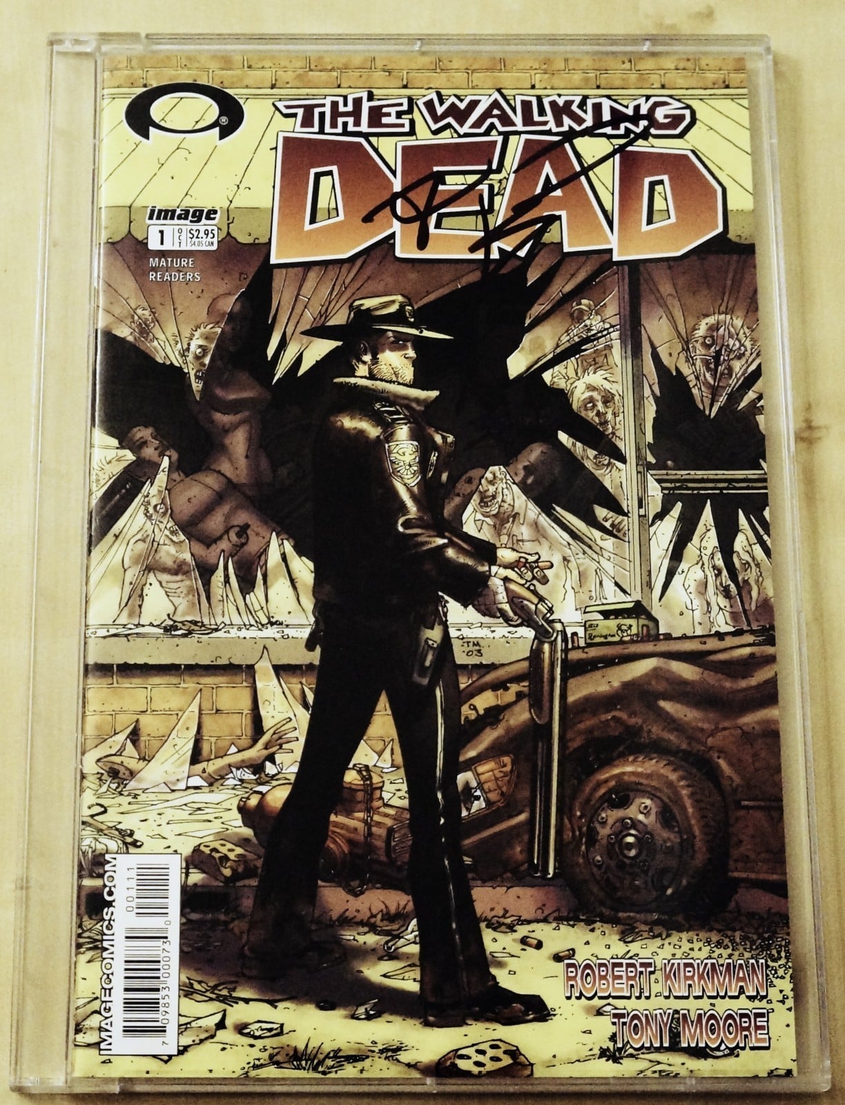 Comic book cover of “The Walking Dead” shows a man in a sheriff’s uniform with a gun, standing by a wrecked car, surrounded by zombies behind glass. Title and author names are visible at the top and bottom.