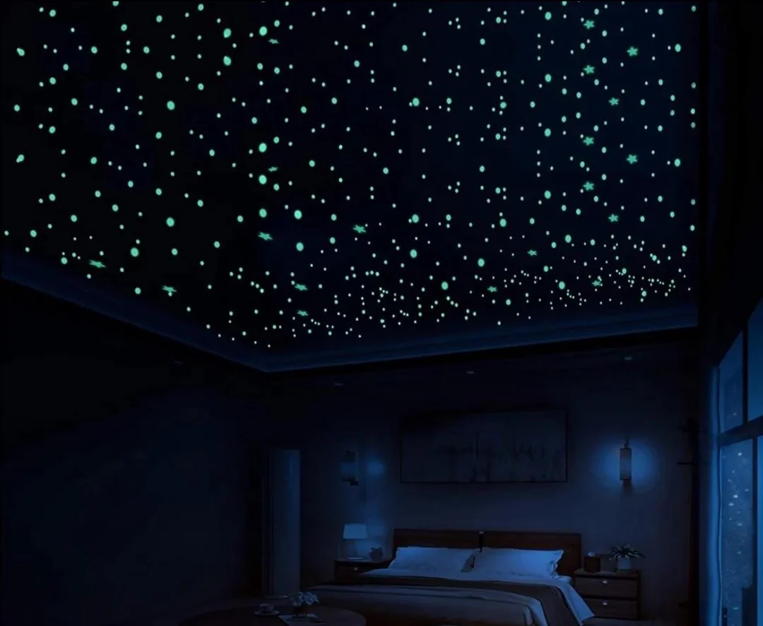 A dark bedroom with a ceiling covered in glowing stars, resembling a night sky. The bed, lamps, and a window with curtains are dimly visible, creating a calm, cozy atmosphere.
