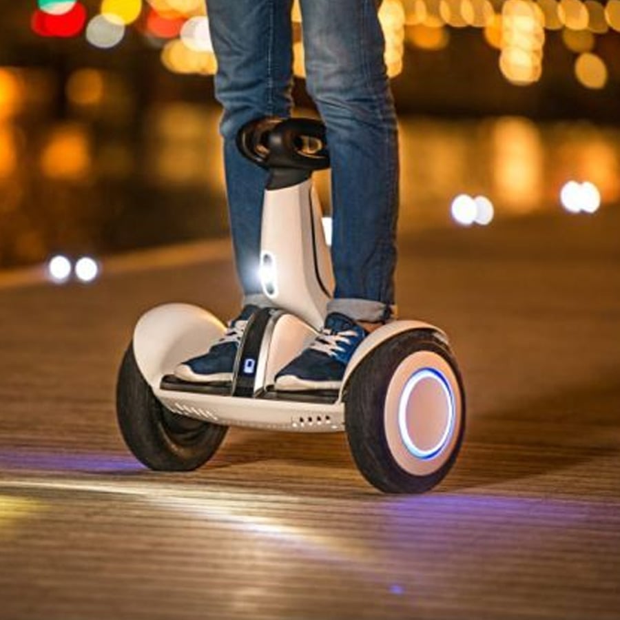 Person standing on a white self-balancing electric scooter at night on a wooden path, with city lights blurred in the background. Only the lower half of the person is visible.