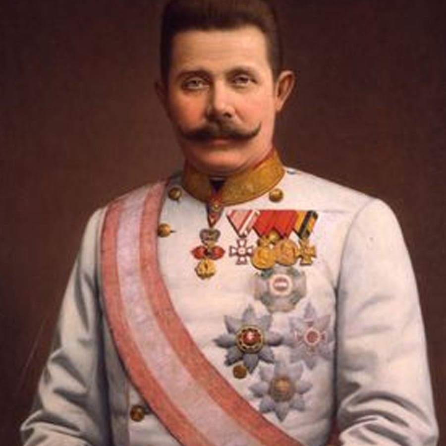 A man in a white military uniform with medals and a sash, dark hair, and a prominent mustache, poses against a plain brown background.