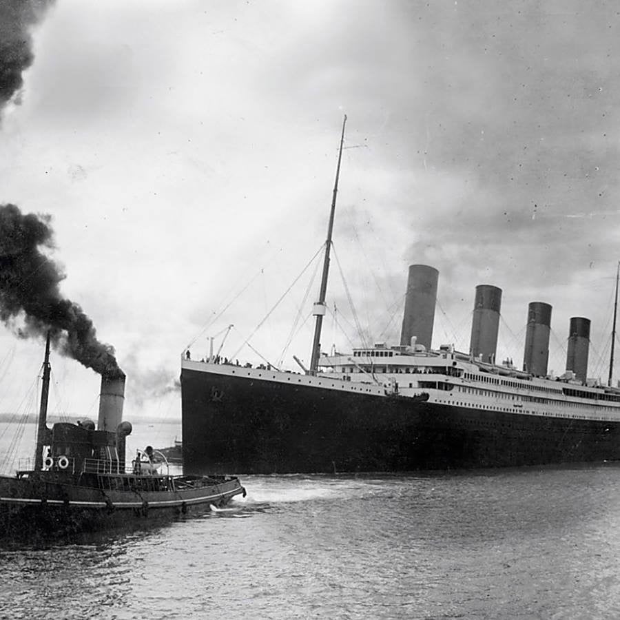 Large, early 20th-century ocean liner with four smokestacks sails near a smaller steam-powered tugboat emitting dark smoke, both on calm water under a cloudy sky.