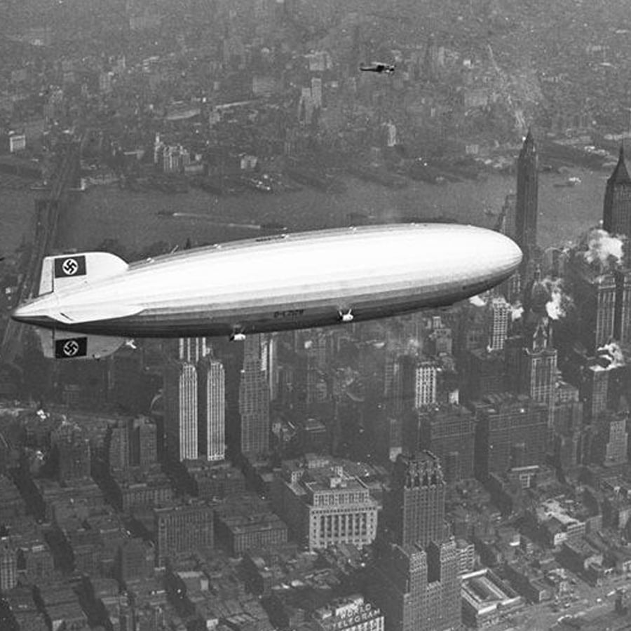 A large zeppelin with swastika symbols on its tail and fin flies over New York City skyscrapers in a black-and-white photo, with the Empire State Building and a river visible below.