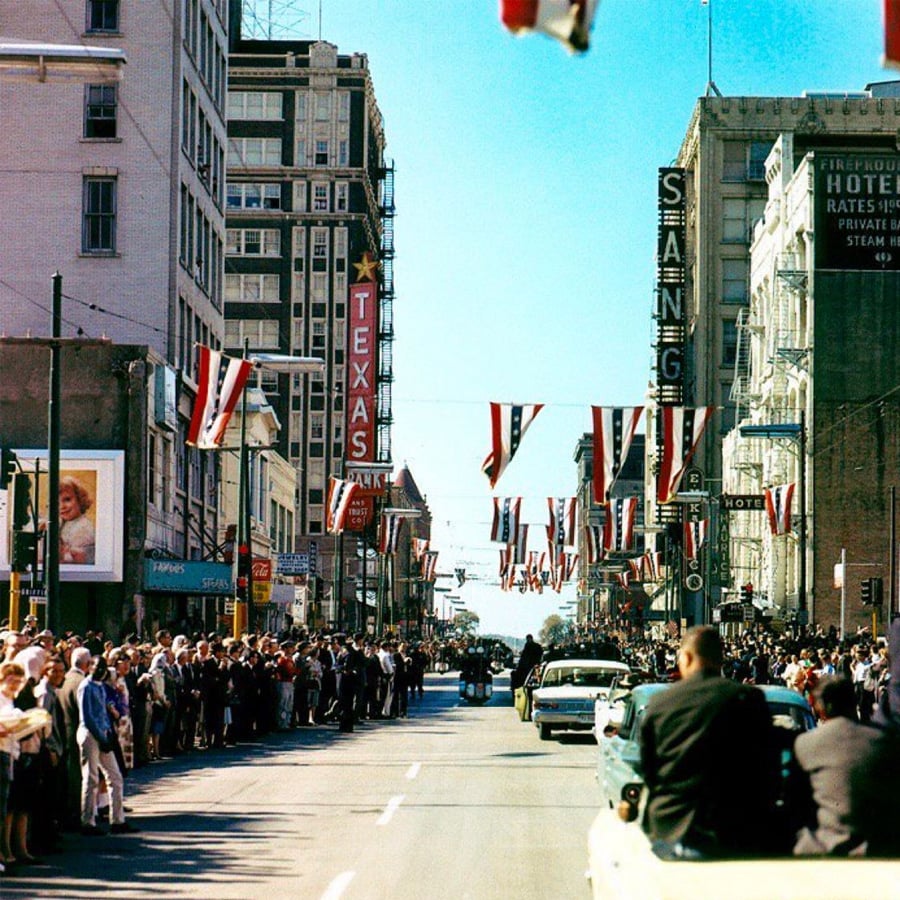 A motorcade drives down a city street lined with crowds and US flags. Tall buildings, including one with a “TEXAS” sign, are visible under a clear sky.
