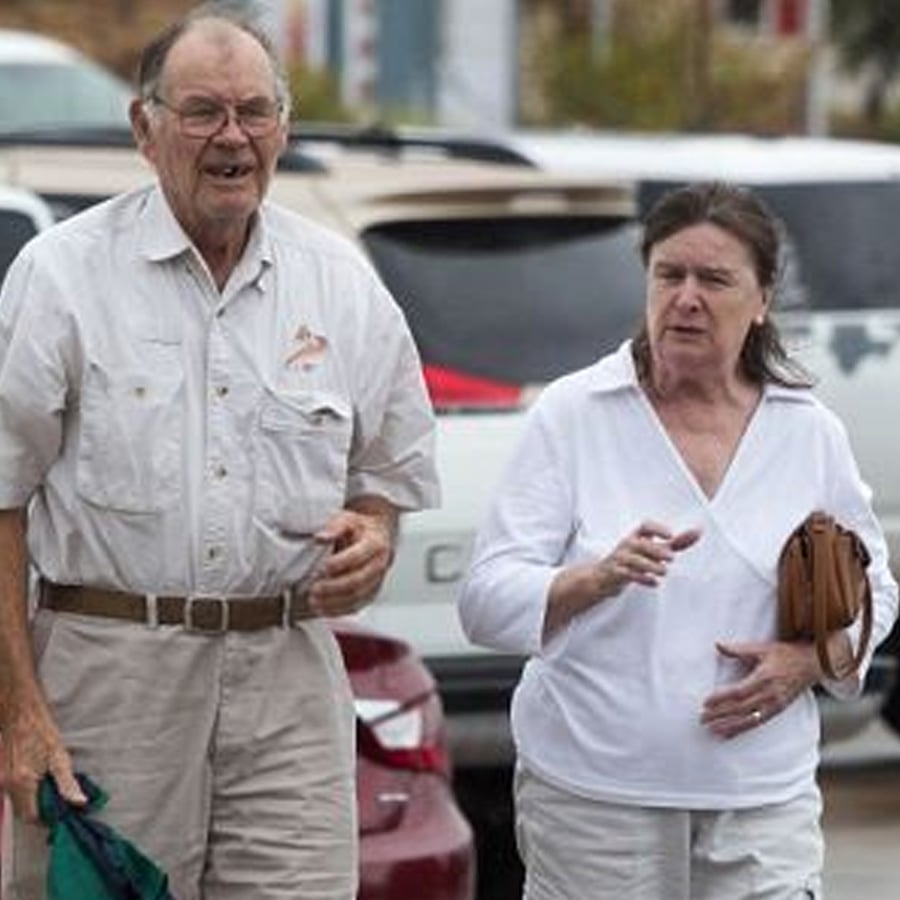An older man and woman walk outside near parked cars. The man wears glasses, a light shirt, and khaki pants. The woman wears a white shirt and holds a brown purse.