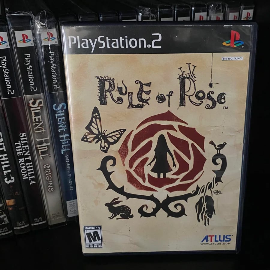 A PlayStation 2 game case for “Rule of Rose” is shown on a shelf next to several Silent Hill games. The cover features a silhouette of a girl inside a large rose, with animals and a butterfly around it.