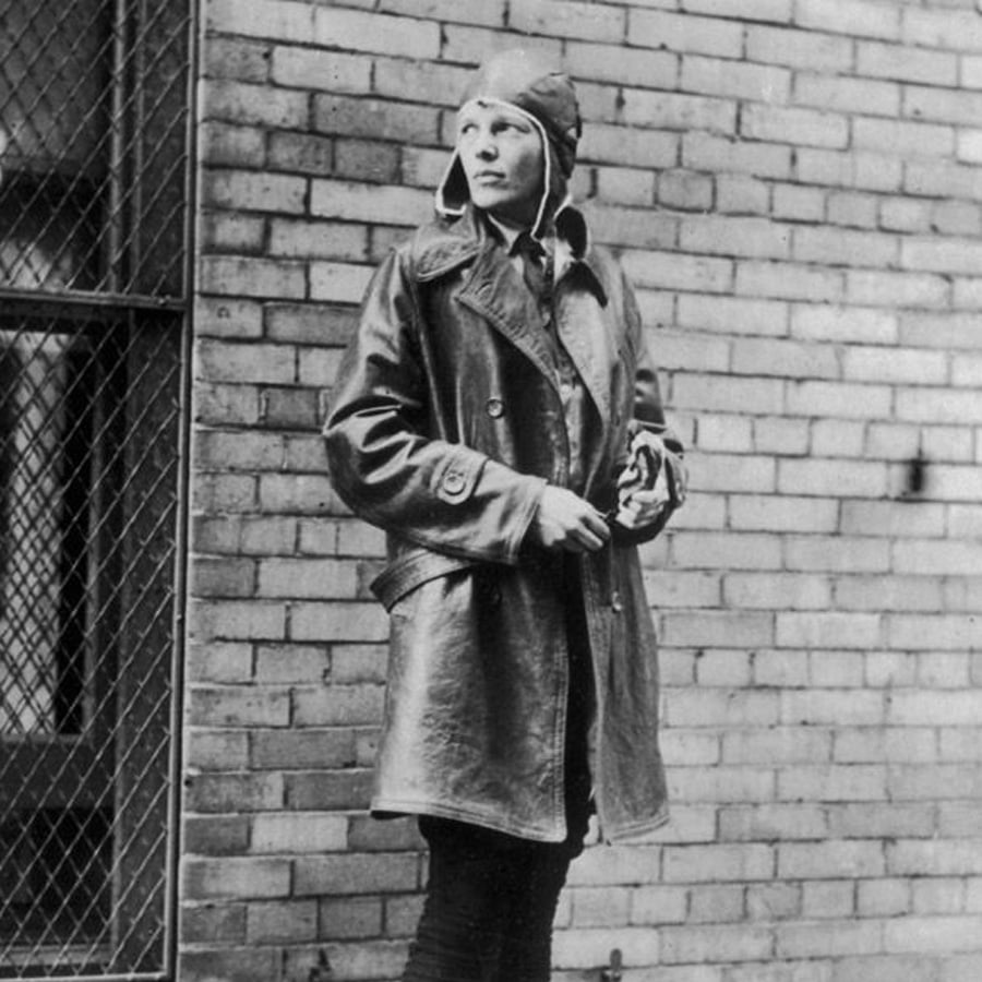 Person wearing a leather coat and aviator cap stands in front of a brick wall, looking to the side with hands clasped, next to a window with a wire mesh covering. Black and white photograph.