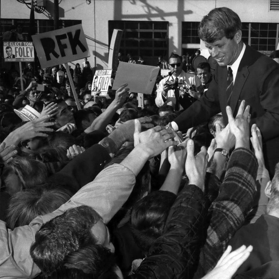 A man in a suit stands above a crowd, shaking hands with people reaching toward him. Signs in the crowd read “RFK” and “Reelect Tom Foley.” The scene appears energetic and enthusiastic.