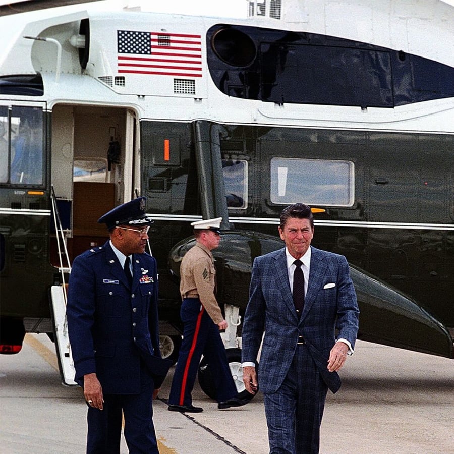 A man in a blue suit walks beside a uniformed Air Force officer near a green helicopter with a U.S. flag, while a Marine stands in the background.