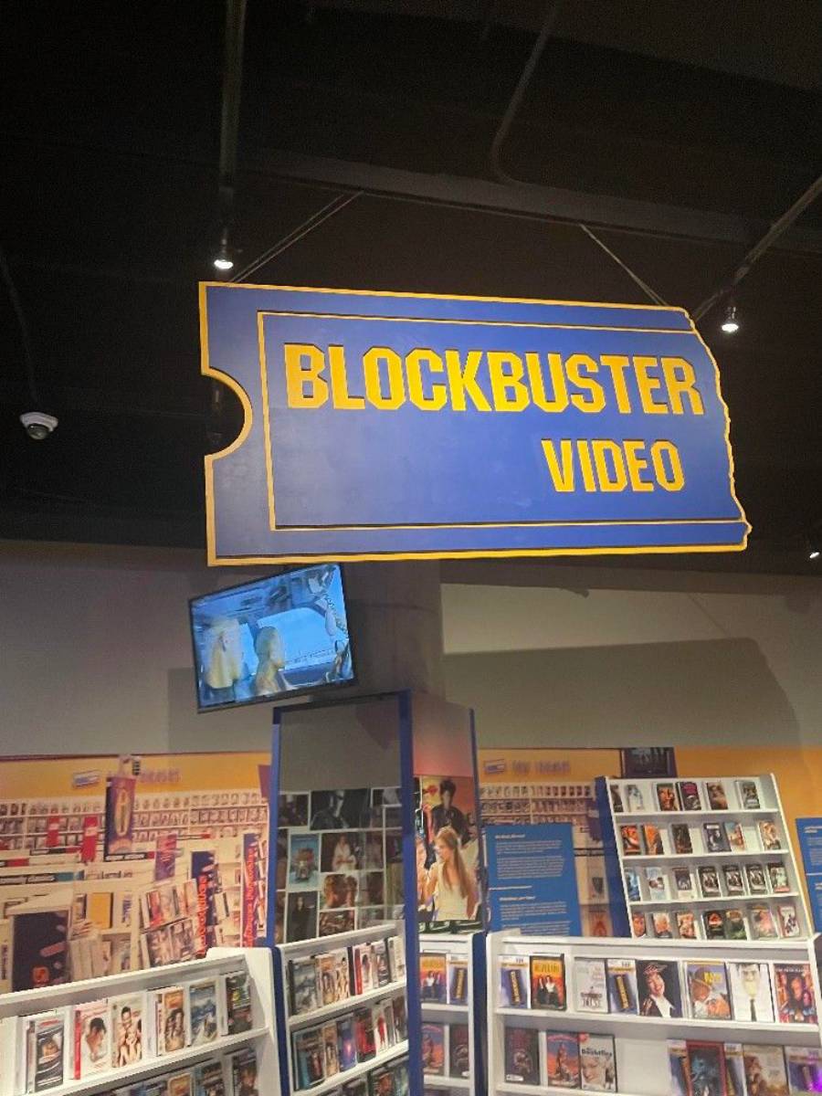 A large blue and yellow “Blockbuster Video” sign hangs above shelves filled with DVDs and VHS tapes, with a TV screen showing a movie scene in the background.