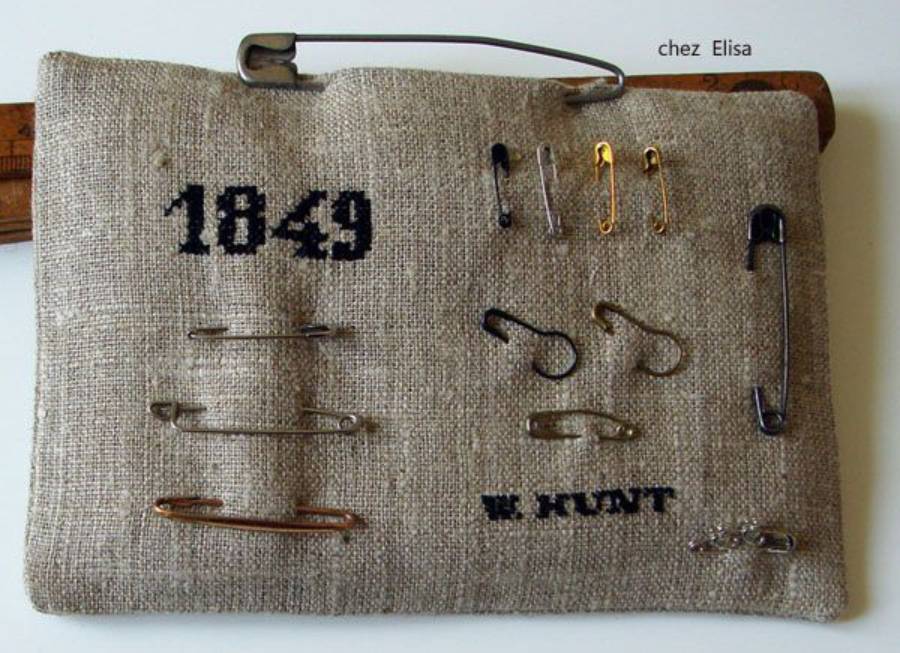 A beige fabric rectangle displays various sizes of safety pins and black embroidered text reading “1849” and “W. HUNT” on the surface. A ruler is partly visible behind the fabric.