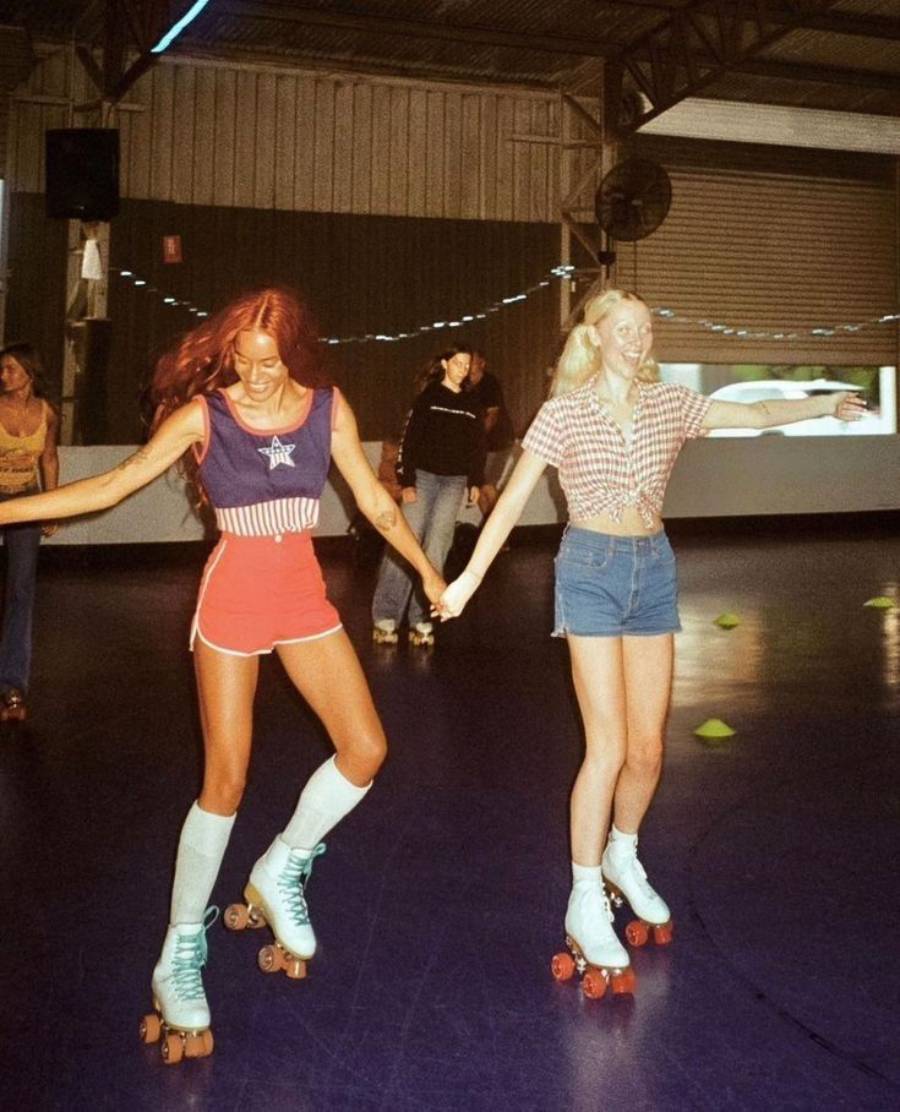 Two young women wearing vintage outfits roller skate and hold hands inside a rink, smiling and looking joyful. The background shows other skaters and a decorated indoor setting.