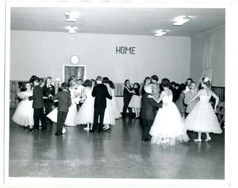 Black and white photo of couples in formal attire dancing in a large hall with “HOME” written on the back wall. Many women wear white dresses and men wear suits.