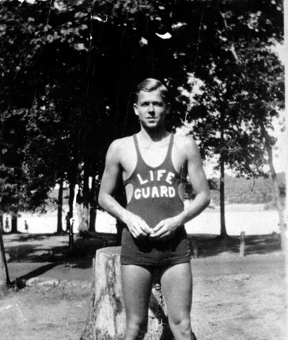 Black and white photo of a young man in a tank top and shorts labeled “LIFE GUARD,” standing outdoors near trees and a lake, with hands clasped in front.