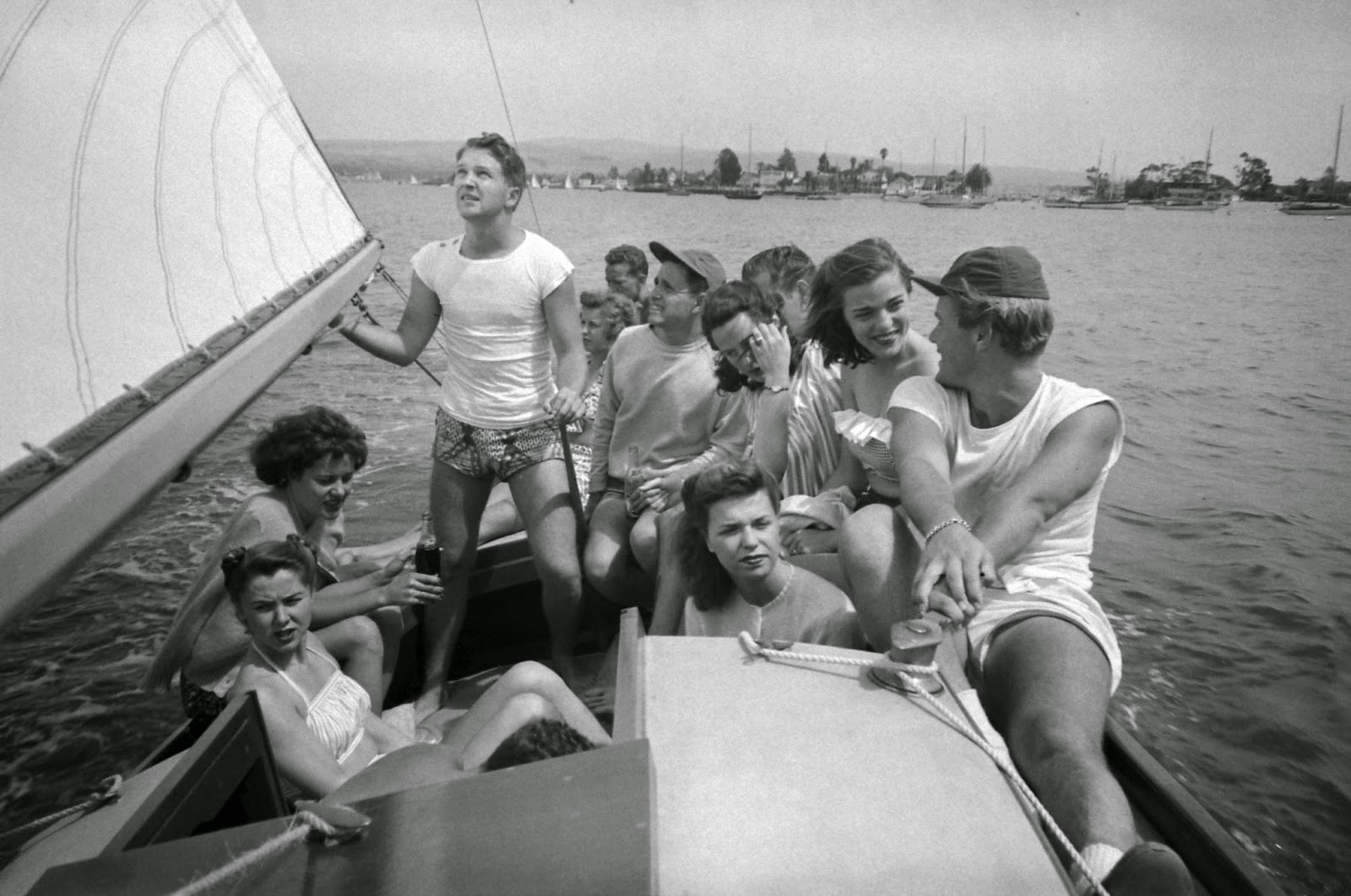 A group of young adults in swimsuits sit and chat on a sailboat out on the water, with one person holding the sail. Other boats and a shoreline are visible in the background.