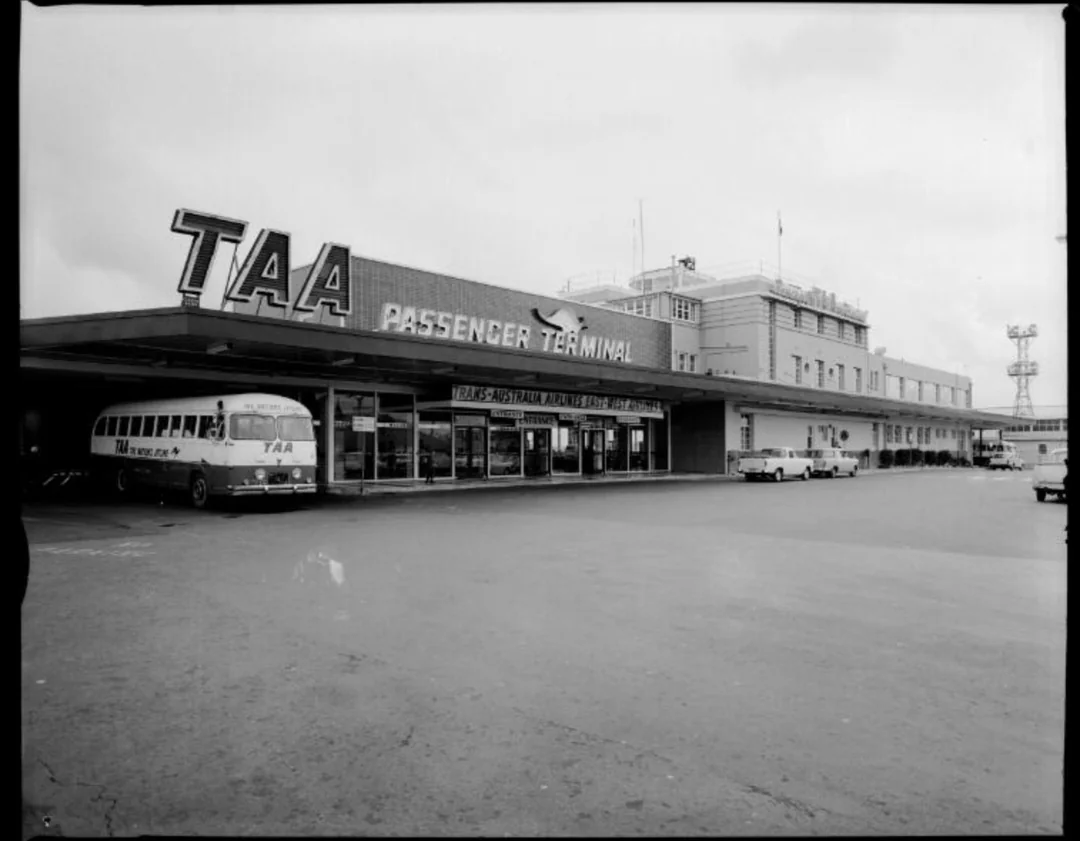 Black and white photo of the TAA Passenger Terminal with a TAA bus parked in front. Large sign on the building, glass entrance doors, a few cars, and a tower are visible.