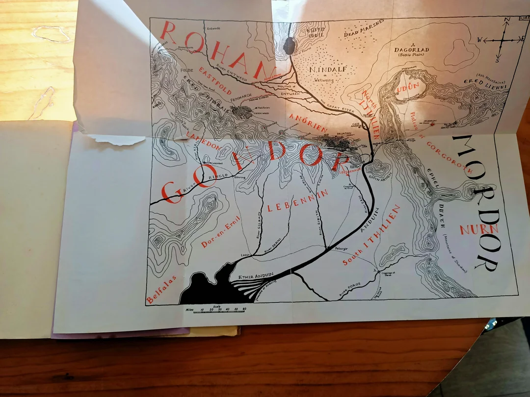 A map from The Lord of the Rings shows regions labeled Rohan, Gondor, and Mordor, with mountains, rivers, and locations like Minas Tirith and Dagorlad, lying open on a wooden surface.