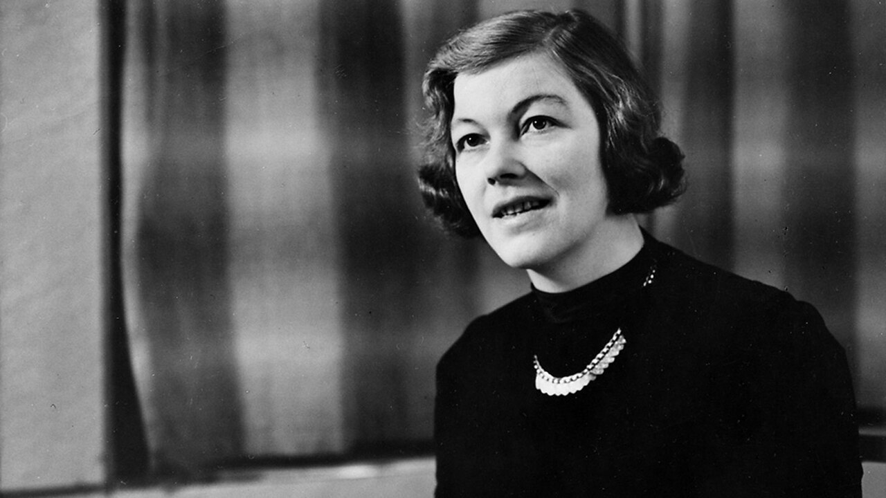 A woman with short, wavy hair and a necklace sits in front of blurred curtains, wearing a dark top and looking slightly to the side with a faint smile. The image is in black and white.