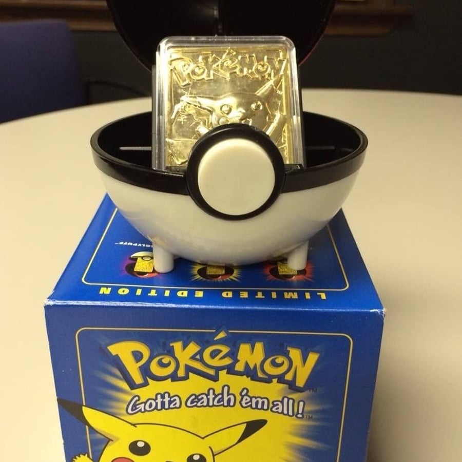 A gold-colored Pokémon card featuring Pikachu is displayed in a black and white Poké Ball case, sitting on a blue “Pokémon: Gotta catch ’em all!” limited edition box with Pikachu artwork.