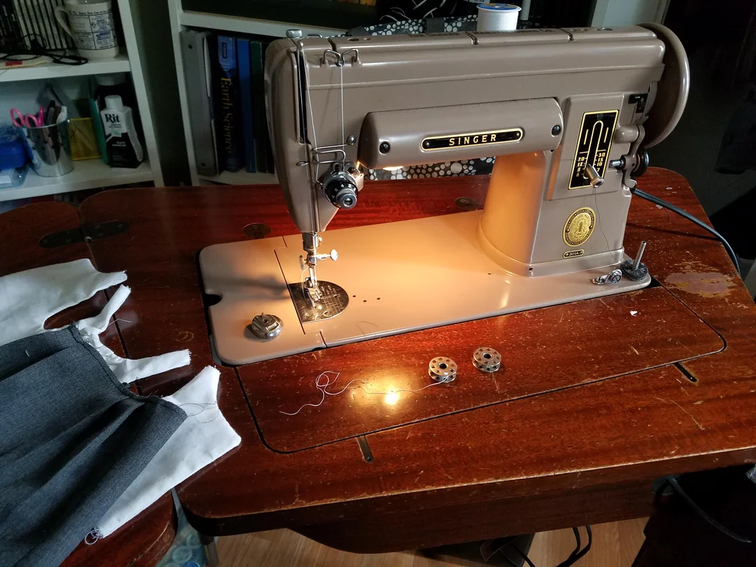 A vintage Singer sewing machine sits on a wooden table with fabric pieces and bobbins nearby, illuminated by a built-in light.