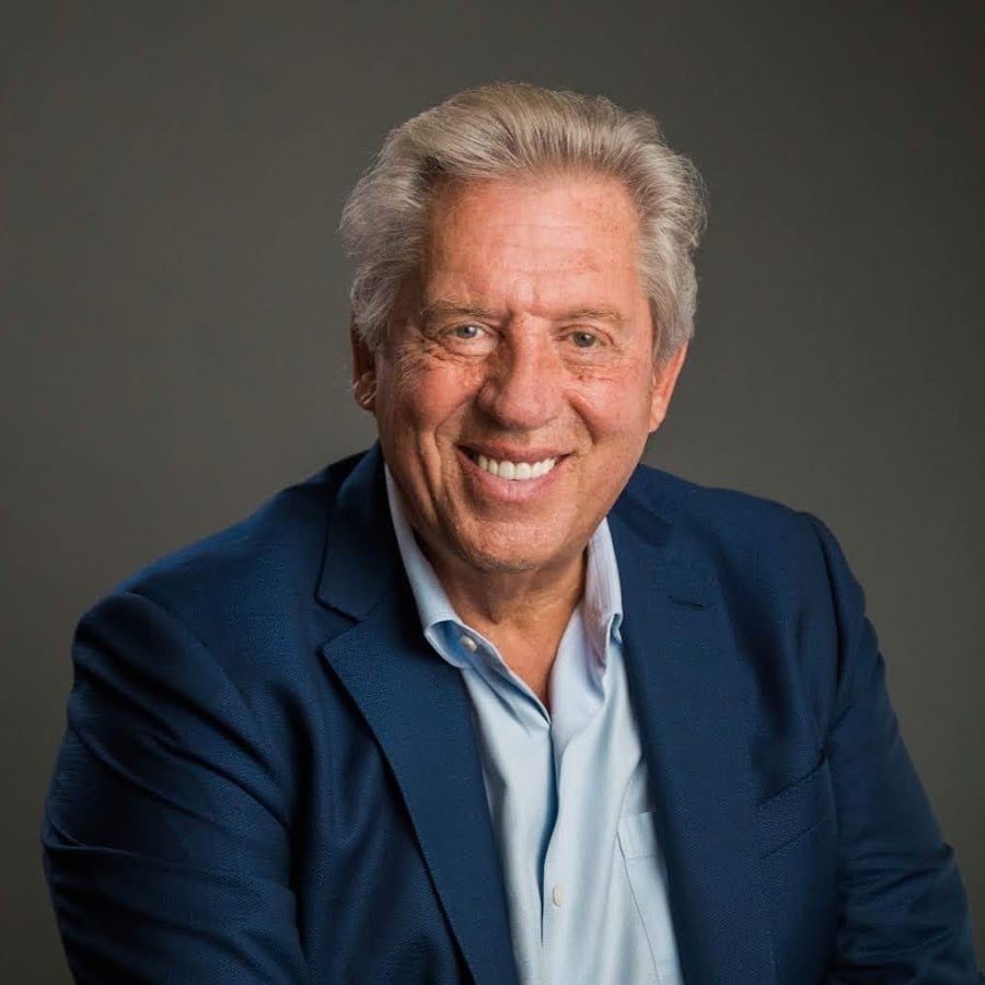 An older man with gray hair smiles warmly at the camera. He is wearing a blue suit jacket over a light blue shirt. The background is plain and dark, keeping the focus on him.