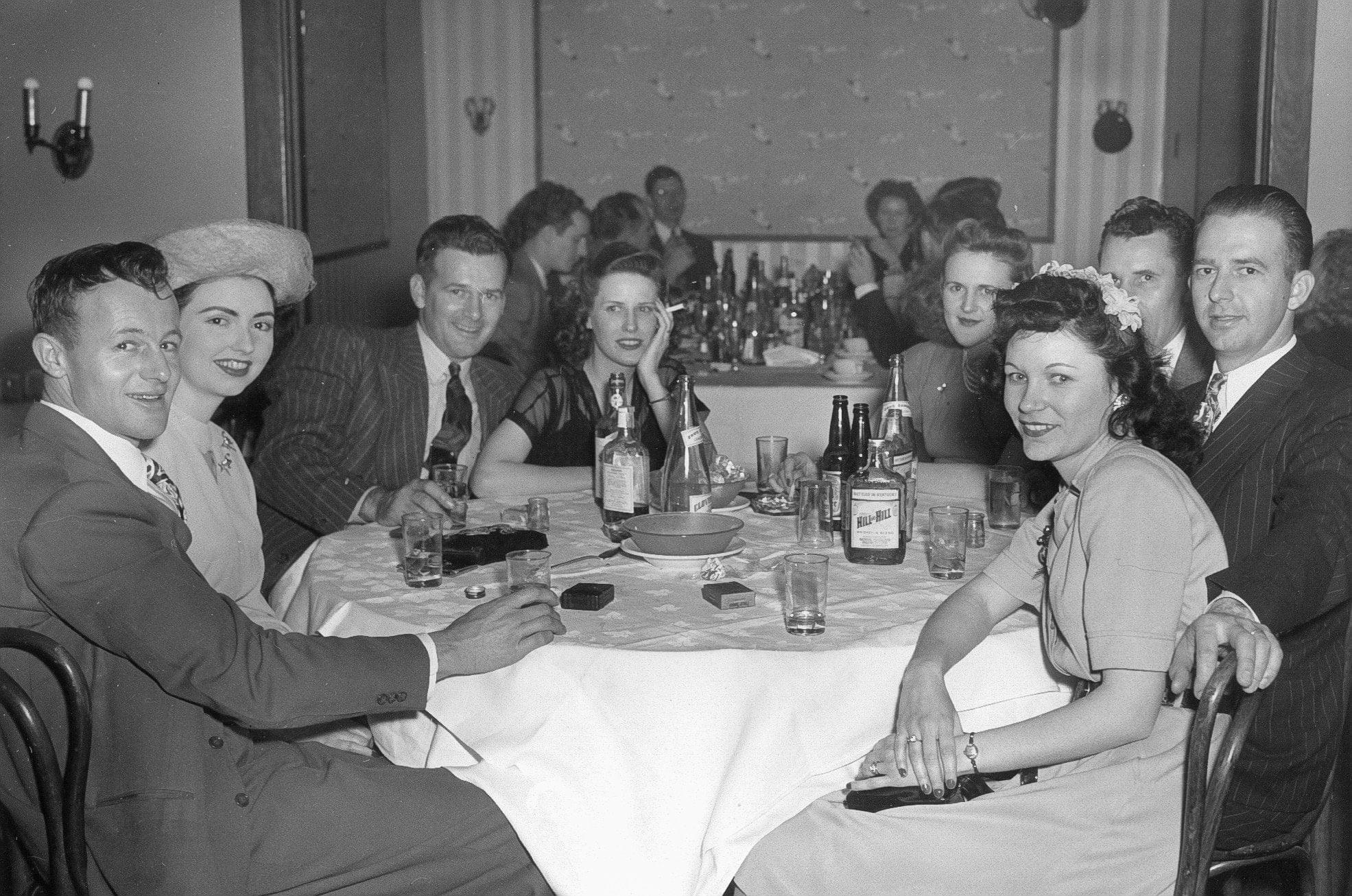 A group of men and women in 1940s attire sit around a table with drinks at a lively social gathering. More people can be seen talking and eating at tables in the background.