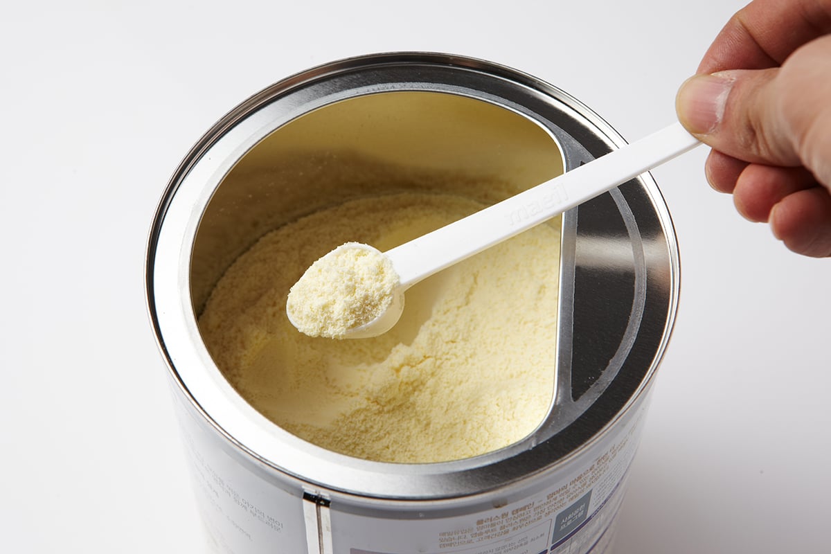 A hand holds a white scoop with powdered formula above an open metal can filled with powdered infant formula on a white background.