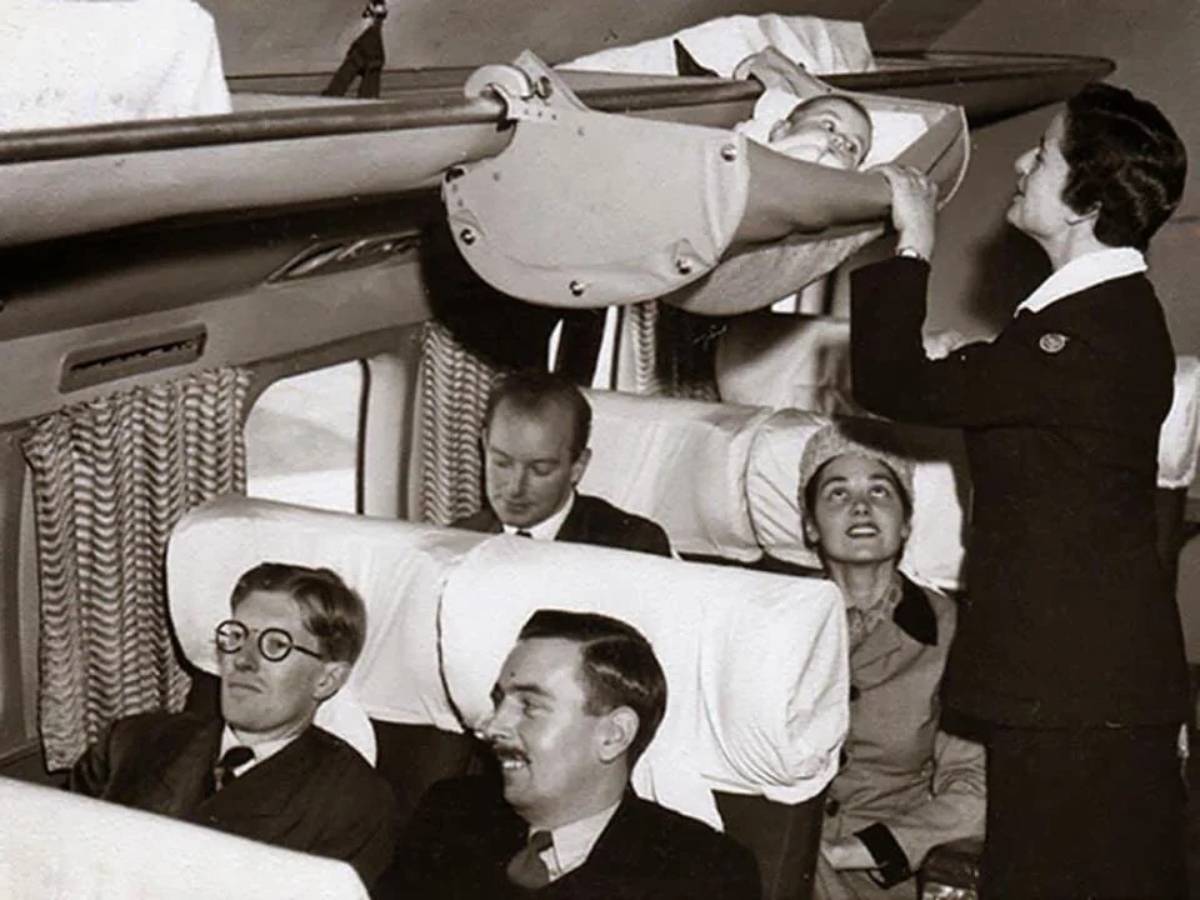 A flight attendant secures a baby in an overhead crib while passengers sit below on an airplane. The black and white photo shows four seated adults watching or relaxing.