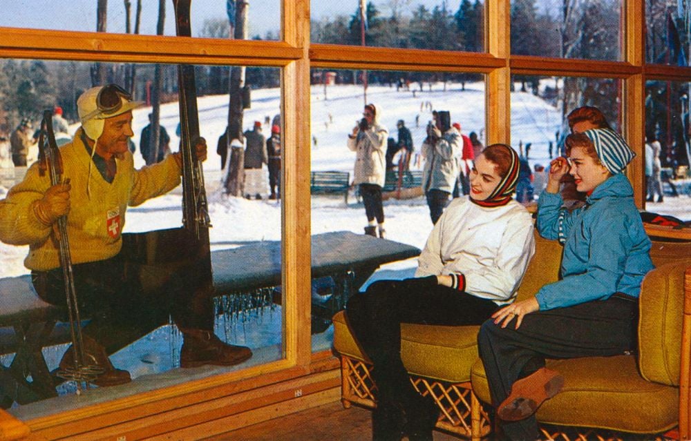 A man in ski gear stands outside a window holding skis, while three women sit on a couch indoors, smiling at him. Snowy outdoor scene with people skiing in the background.