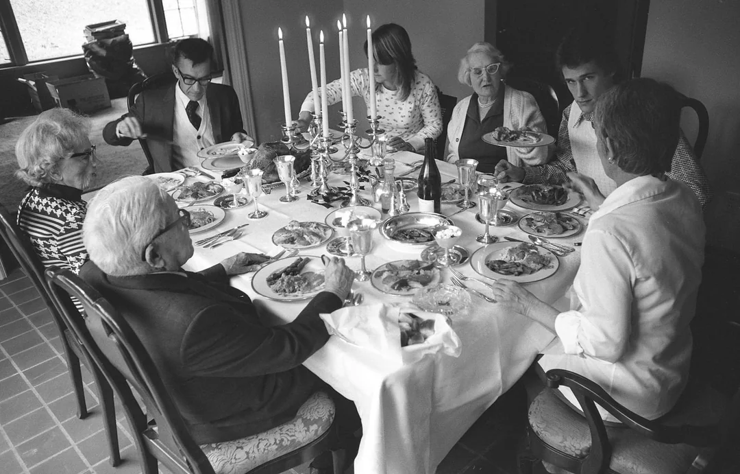 Seven people sit around a dining table set with plates of food, silverware, and tall candles. The group appears to be enjoying a meal together in a well-lit room.