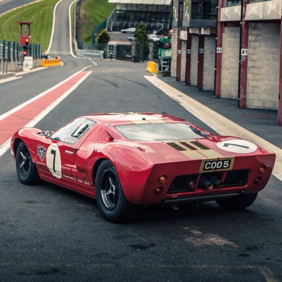 A vintage red race car with the number 7 on the hood is parked on a race track near pit garages, with a curving track visible in the background.