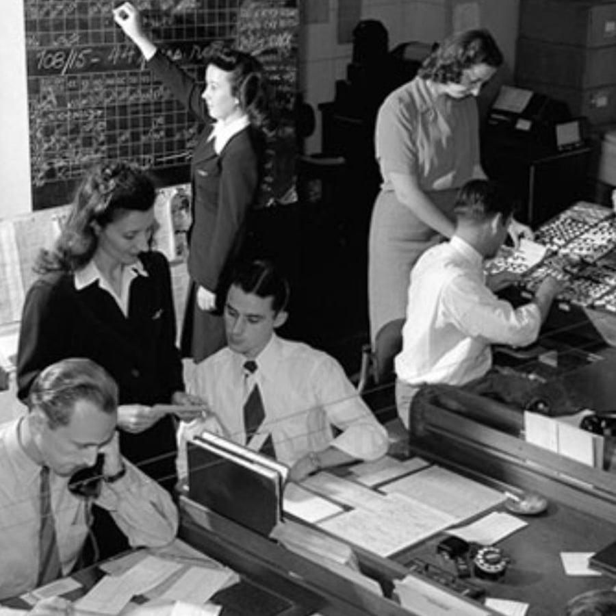 Black-and-white photo of men and women working in an office. Some are seated at desks with papers, while others stand, one writing on a large wall chart. The scene appears busy and organized.