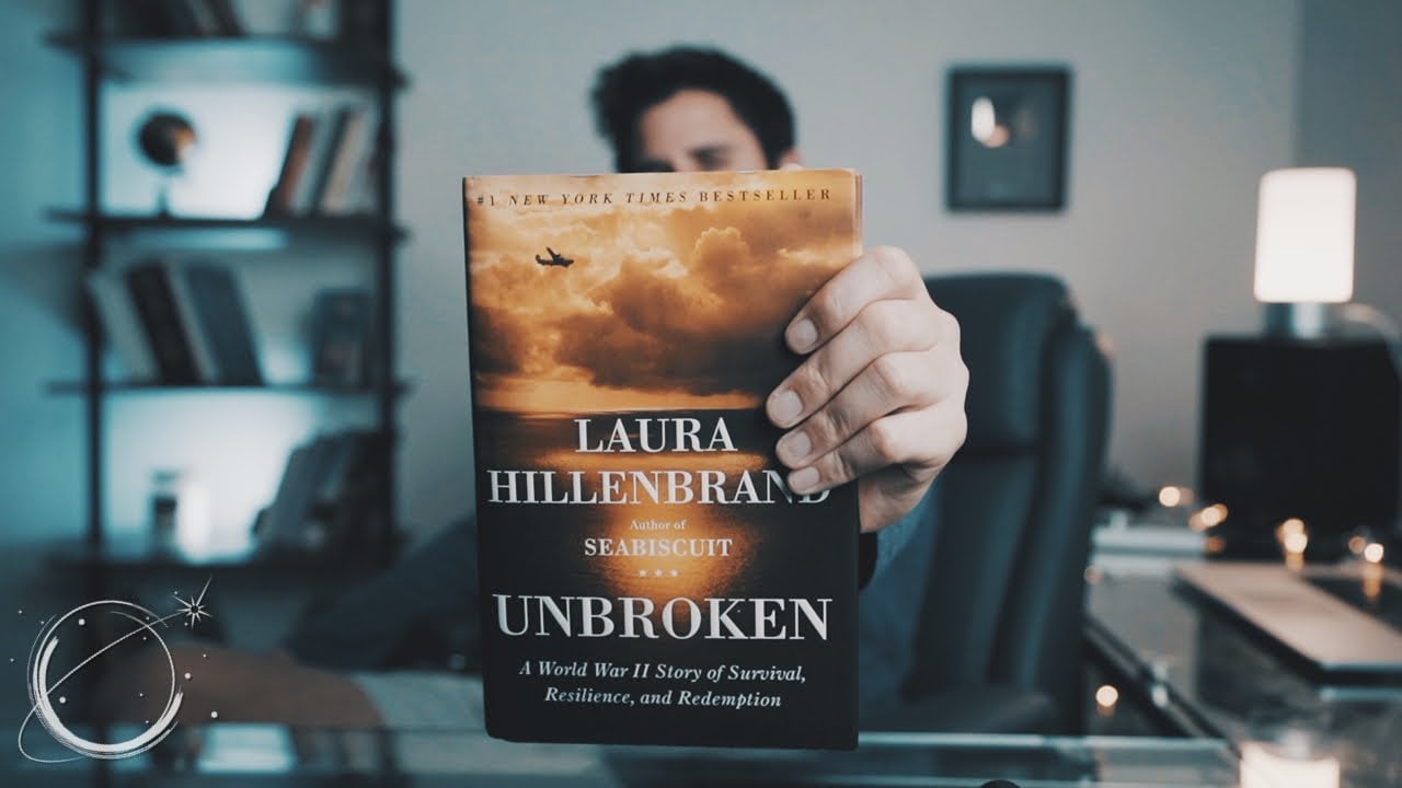 A person holds the book “Unbroken” by Laura Hillenbrand, covering their face, while sitting at a glass desk in a modern, softly lit office or study.
