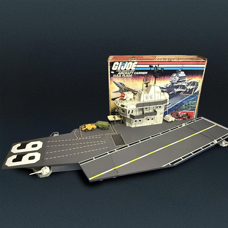GI Joe USS Flagg aircraft carrier playset is assembled on a dark surface, with vehicles on the runway, and its original illustrated box standing behind it.