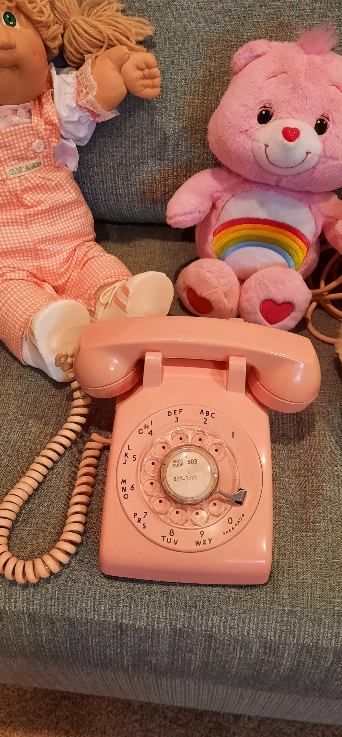 A pink rotary phone sits on a gray couch next to a rag doll in pink overalls and a pink teddy bear with a rainbow on its belly.