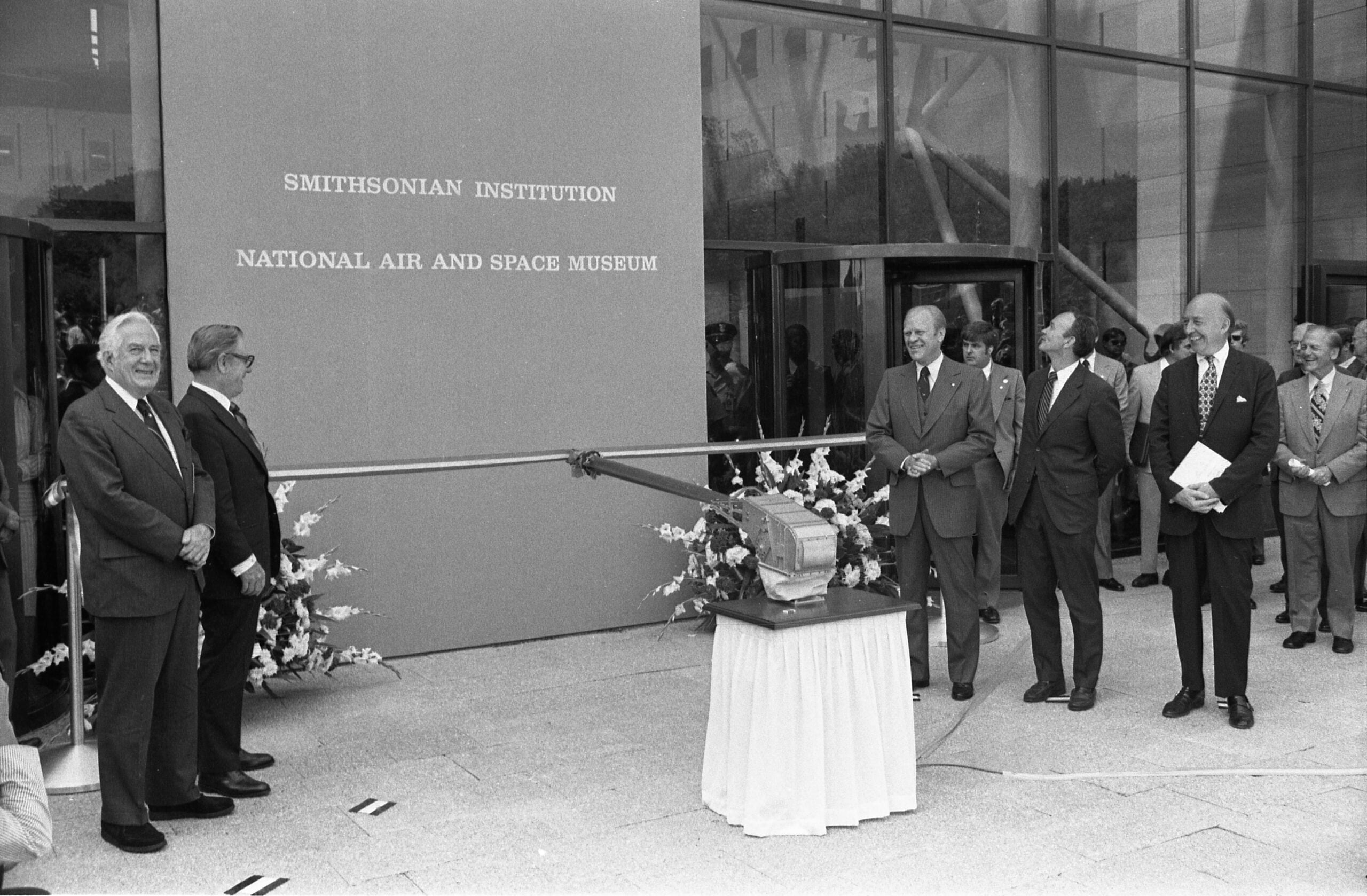 Black-and-white photo of men in suits smiling near the entrance of the Smithsonian Institution National Air and Space Museum, with a ribbon ready to be cut for an opening ceremony.