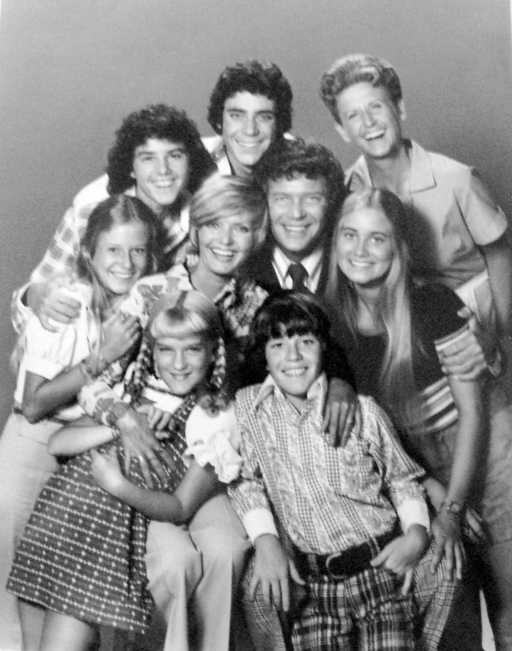 A smiling blended family of nine, including parents, six children, and a housekeeper, pose closely together in a studio portrait, reminiscent of 1970s fashion and style.