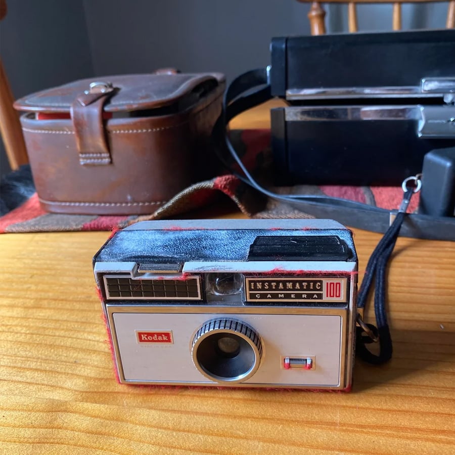 A vintage Kodak Instamatic 100 camera sits on a wooden table, with its brown carrying case and another black case in the background. Warm sunlight streams onto the scene.