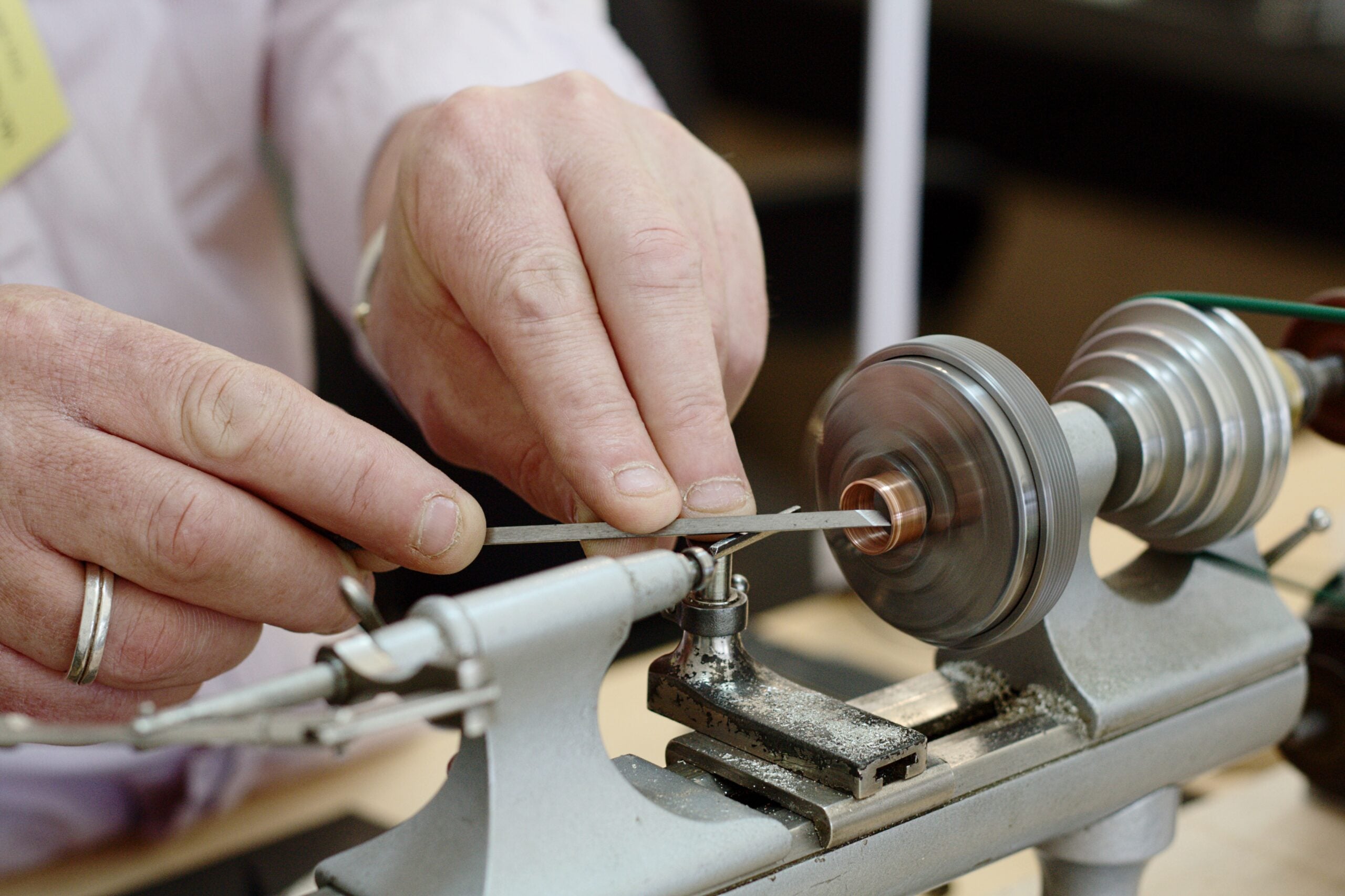 Close-up of hands using a file on a metal piece spinning on a small lathe, demonstrating precise metalworking or machining in a workshop setting.