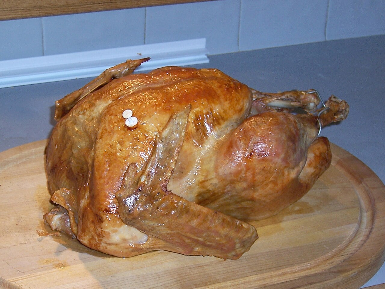 A whole roasted turkey with golden brown skin sits on a wooden cutting board on a kitchen counter.