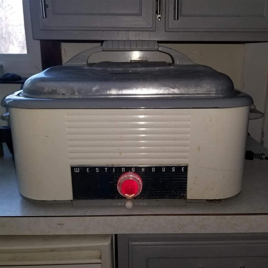 A vintage Westinghouse roaster oven with a metal lid sits on a kitchen counter. The appliance has a white body, ribbed front, and a red knob labeled “Time” and “Temp” below the Westinghouse brand name.