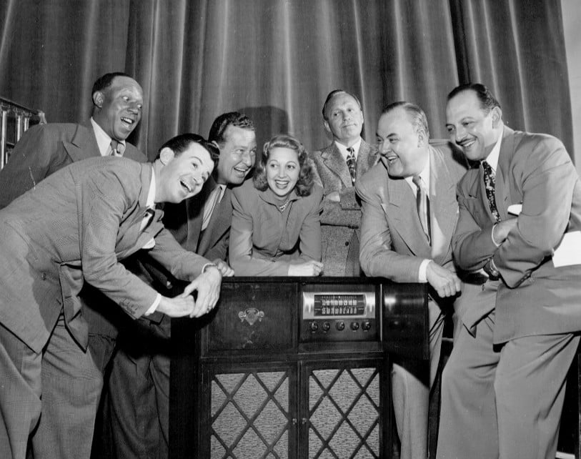 Seven people in suits and dresses smile and gather around a vintage radio, posed in front of a curtain in a black-and-white photo, suggesting a classic mid-20th century setting or broadcast event.