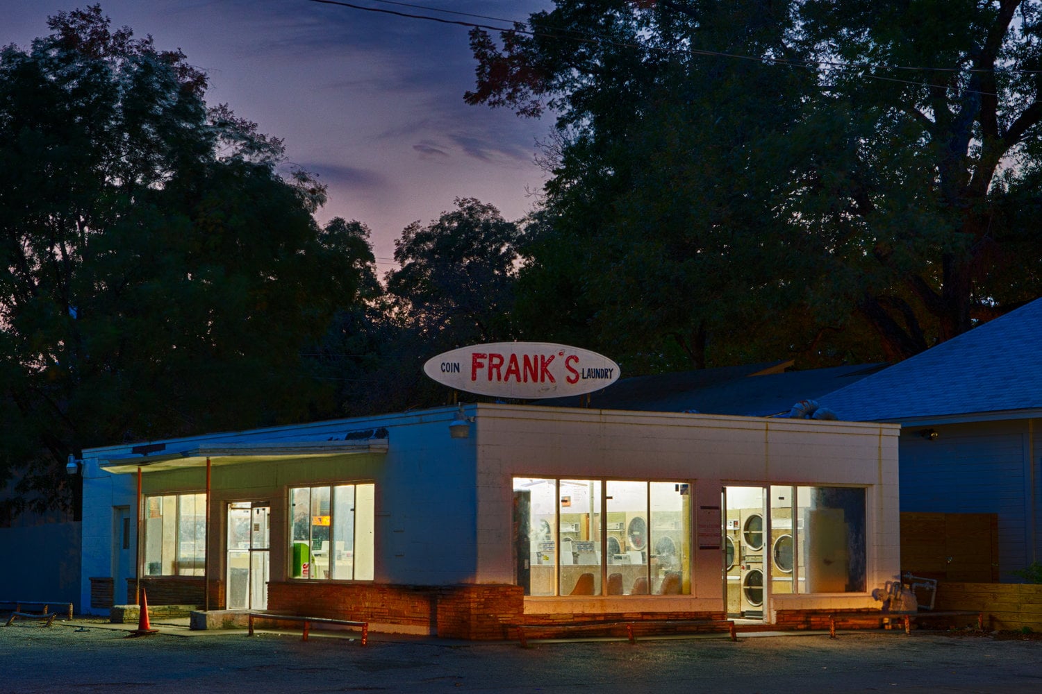 A laundromat named “Frank’s” is lit up at dusk, with large windows showing washing machines inside. The sky is dark blue and trees surround the building.