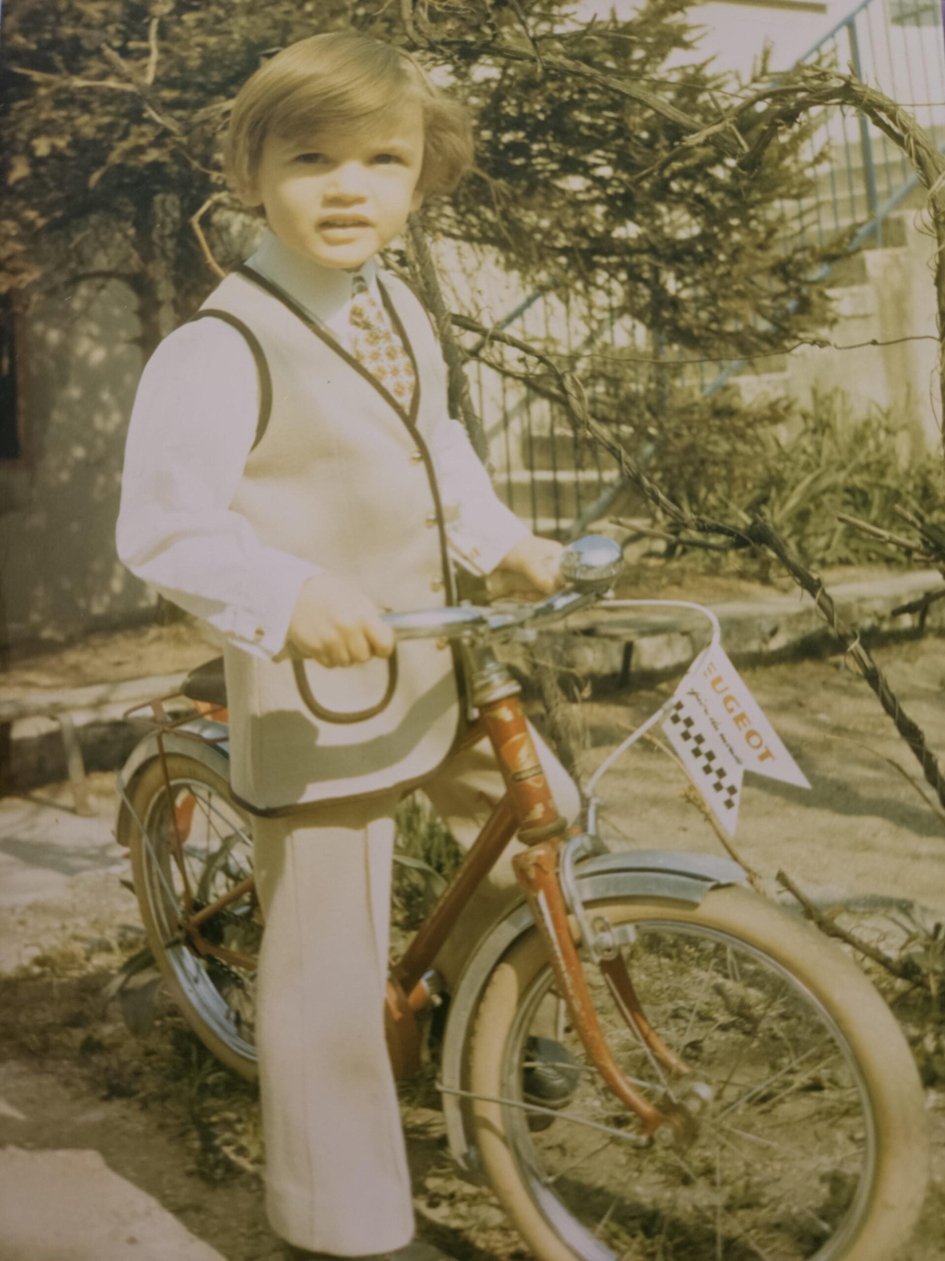 A young child wearing a beige suit and tie stands beside a red bicycle with a “Peugeot” flag, outdoors on a sunny day, with trees and a building in the background.