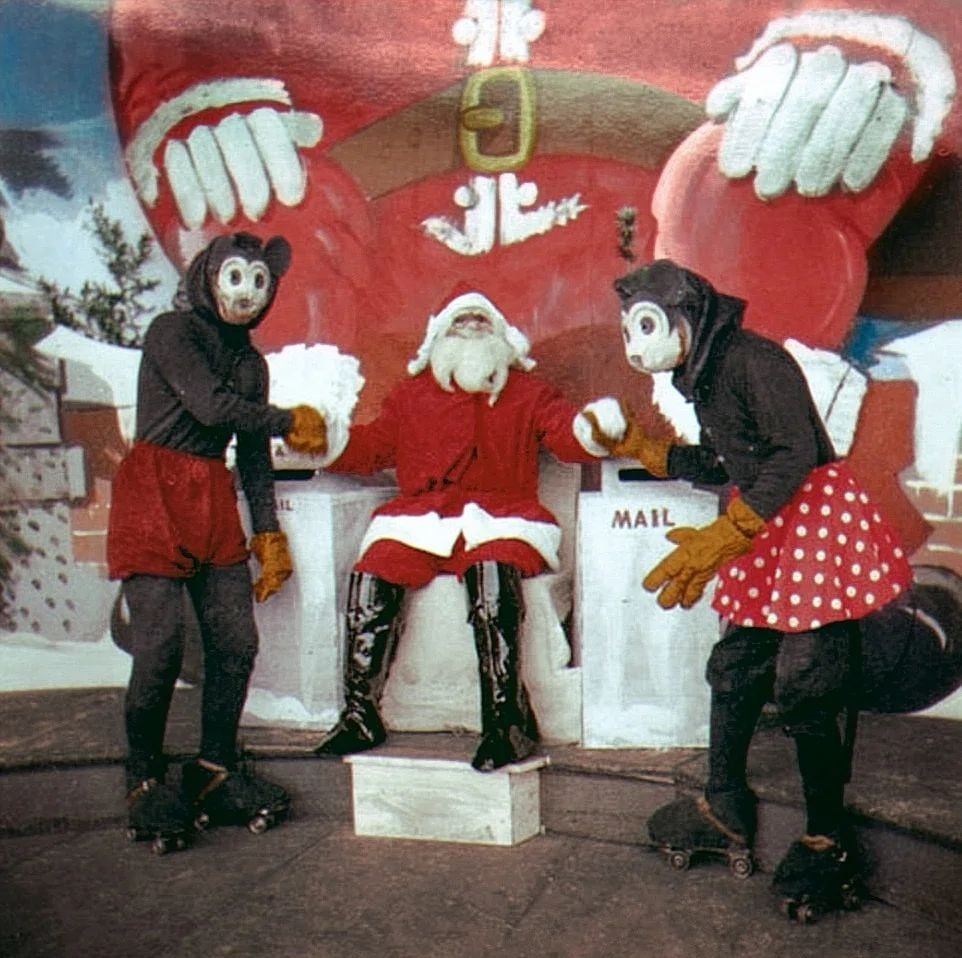Three people pose in front of a giant Santa display: one dressed as Santa and two in vintage cartoon character costumes with mouse heads, gloves, and roller skates. There are two mailboxes labeled “MAIL” on either side.