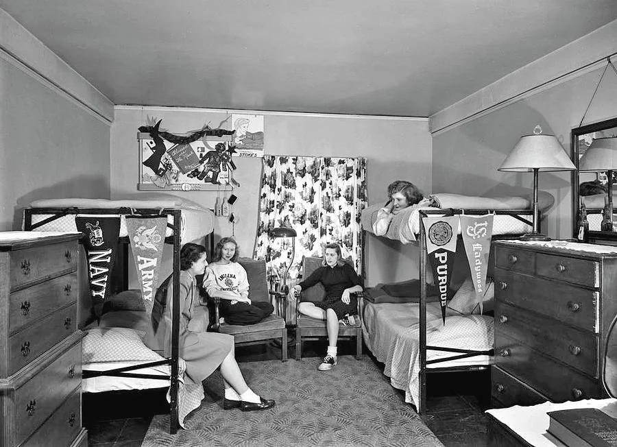 Four young women sit and chat in a neatly arranged dorm room with two sets of bunk beds, college pennants on the walls, and a window with patterned curtains.