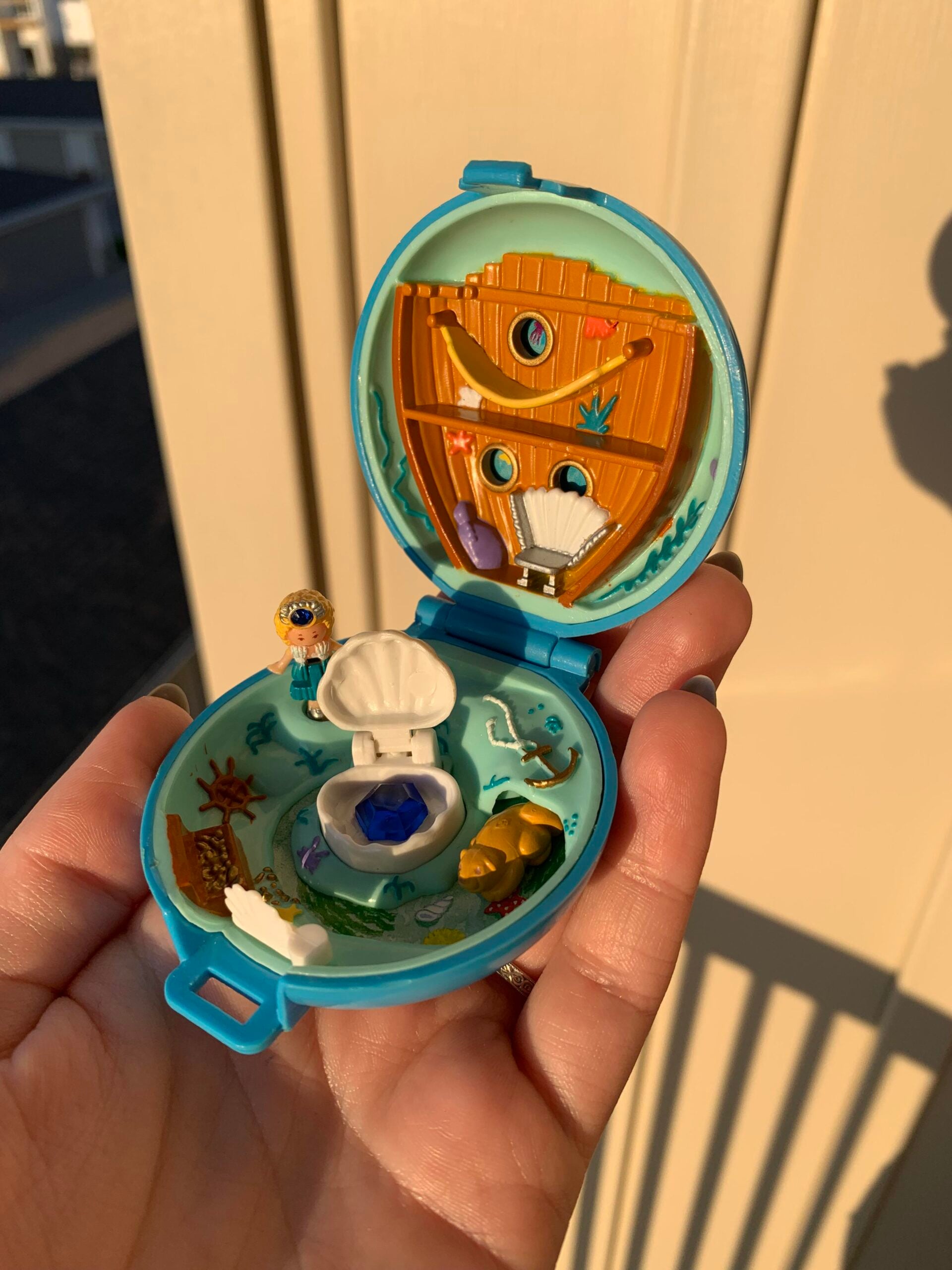 A hand holds an open blue seashell-shaped toy, revealing a tiny underwater scene inside with a small doll, a shell, and sea-themed decorations, all brightly lit by sunlight.