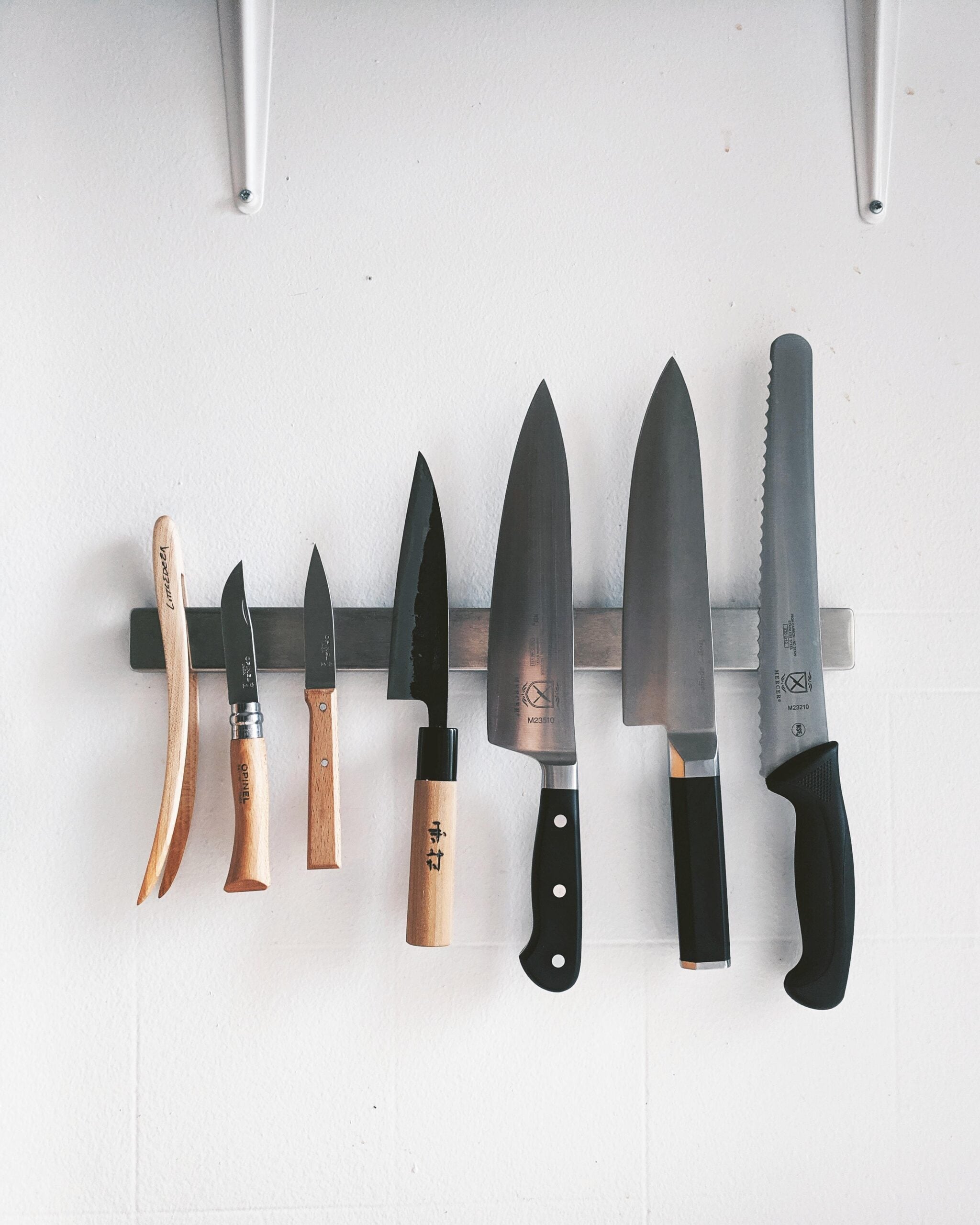 Seven kitchen knives with wooden and black handles are arranged on a magnetic strip attached to a white wall. The knives vary in size, including a bread knife on the far right.