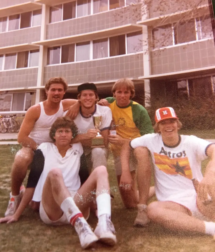 Five young men sit and kneel on grass in front of a building. One holds a trophy, another wears an Astros shirt and cap. They appear happy and relaxed, posing for the photo.